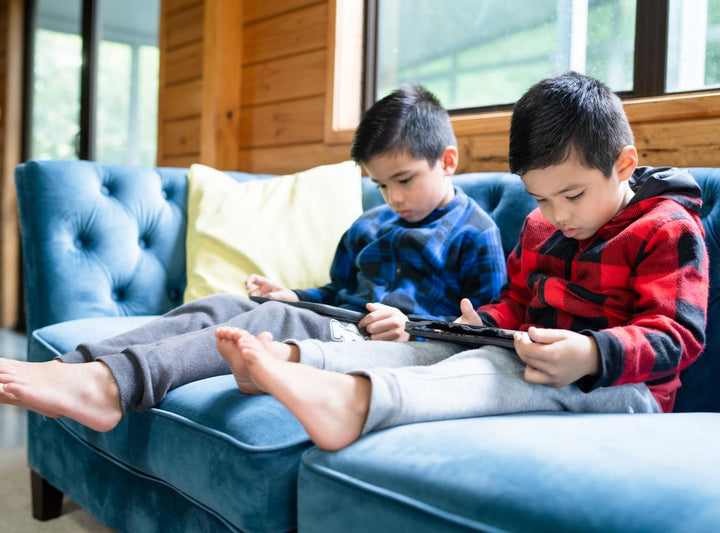 There is a pair of boys on a blue couch on iPads.
